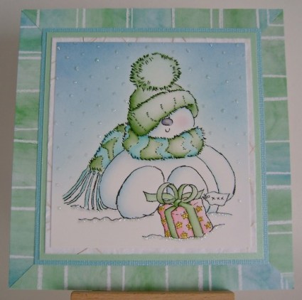 snowgirl wearing green hat with blue and green scarf on striped bue and green framed card