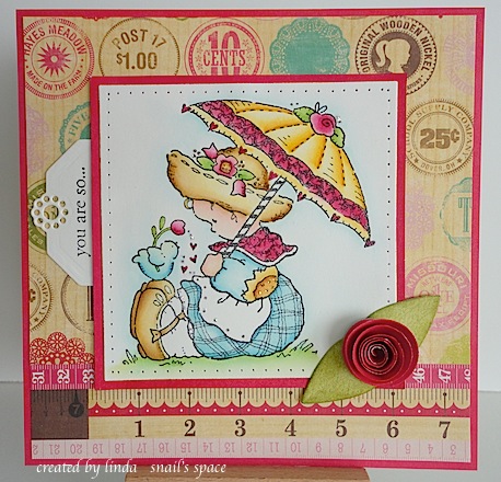 card with girl sitting on ground holding an umbrella with a bird standing on her shoe holding a pink tulip for her with hearts gently falling next to it